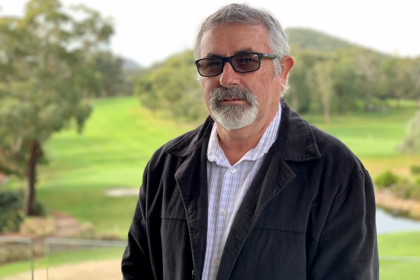 Grey-haired man standing in front of golf course, wearing sunglasses, a dark jacket and open-necked white shirt