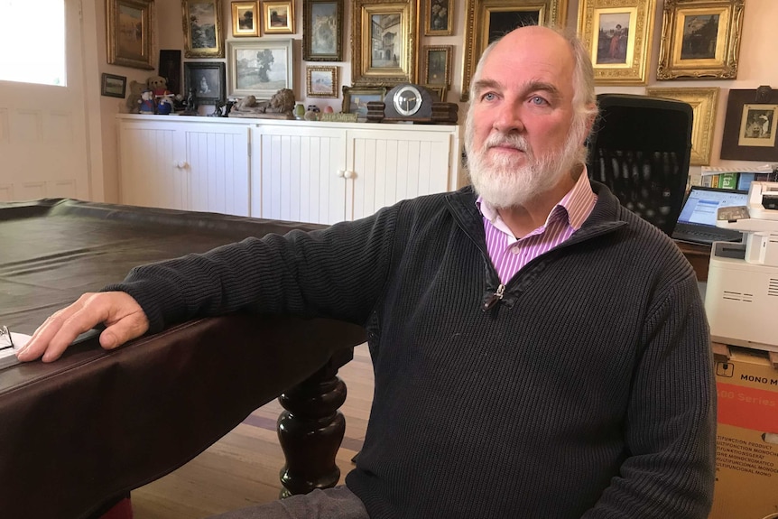 An older gentleman with a grey beard, a bald head and wearing a corduroy jumper sits and stares off into the distance.