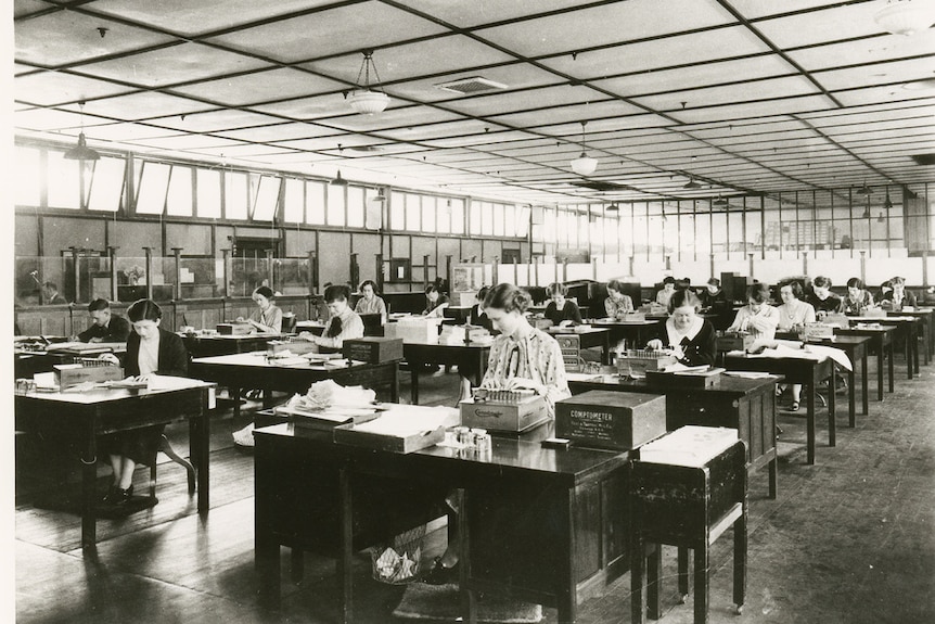 A black and white photo of a large, open office with people working at wooden desks.
