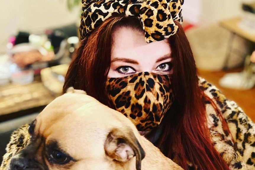 A woman holds her dog wearing a leopard print outfit and mask.