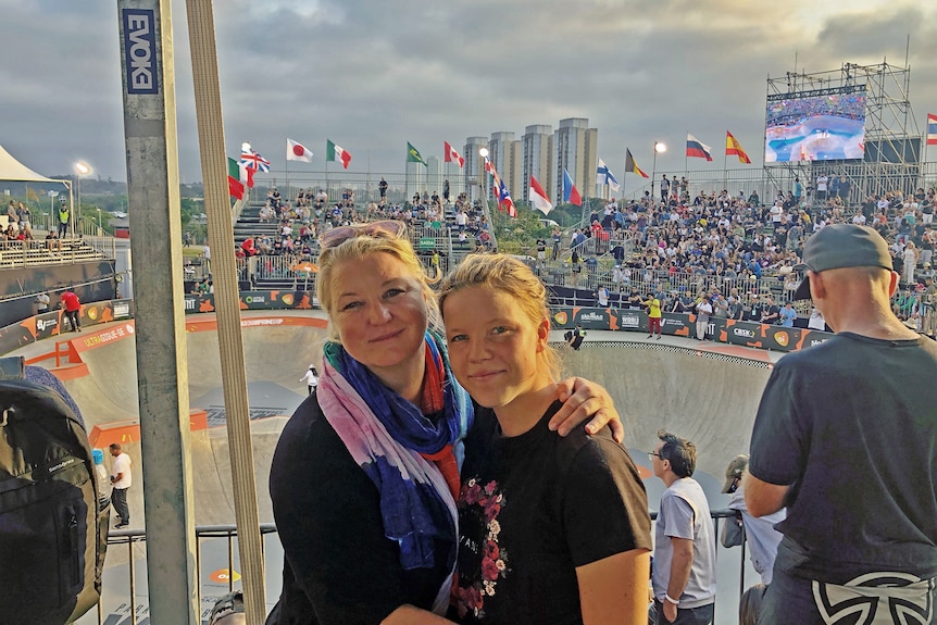 Exterior scene mother and daughter at skateboard competition with bowl and crowd in background