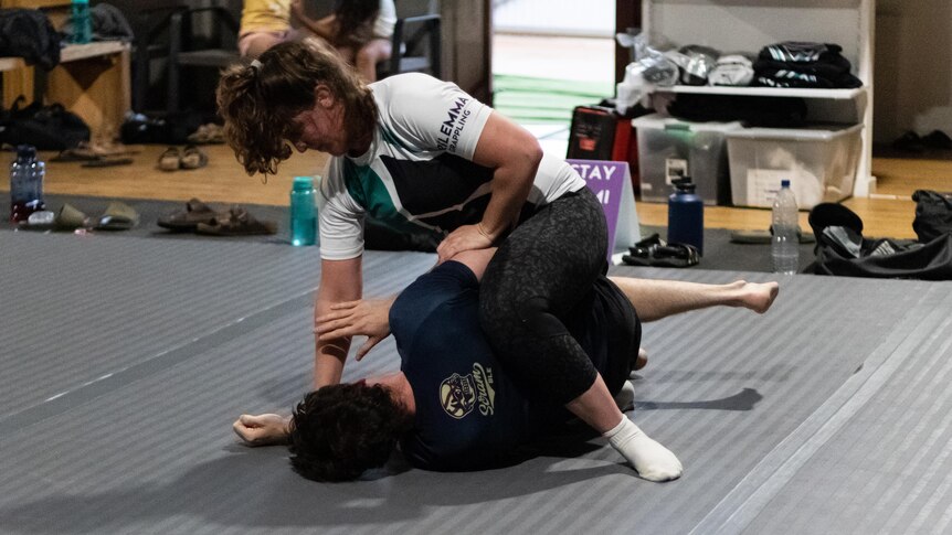 A woman in a white t-shirt wrestles with another participant on a black mat indoors.