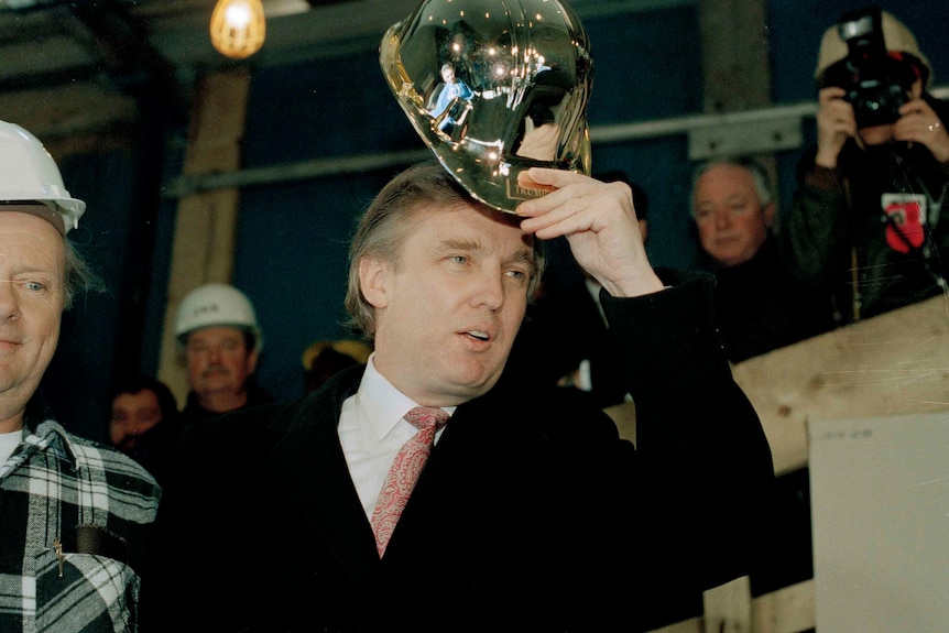 A young Donald Trump tips a gold hard hat as he walks past several construction workers