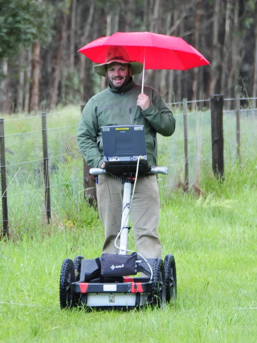 A man with a red umbrella pushes a lawnmower-looking device.