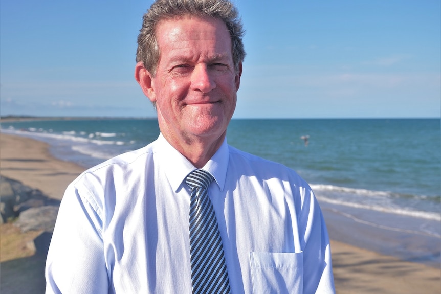 A man smiles at the camera, light blue, long sleeve button up shirt, stripy tie, beach in the background.