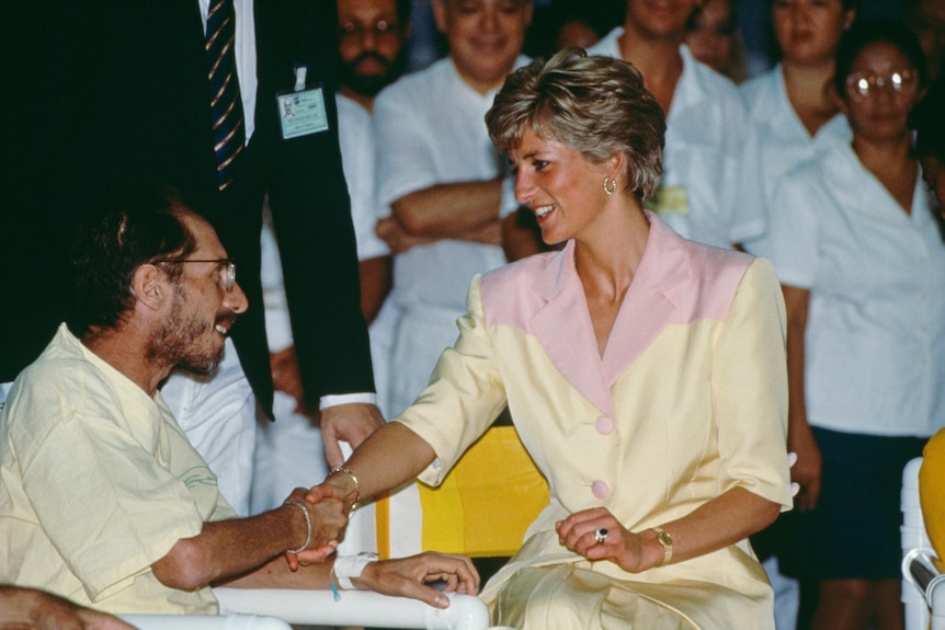 Princess Diana, in pale yellow and pink jacket, smiles and shakes hands with a man, also smiling. Both are seated. 