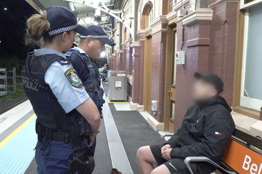 Two police officers speak with a person sitting on a train station bench