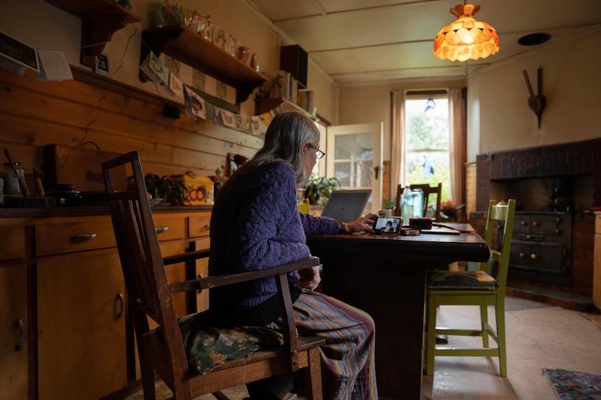 A woman sits at her kitchen table, on a Zoom call on her phone.