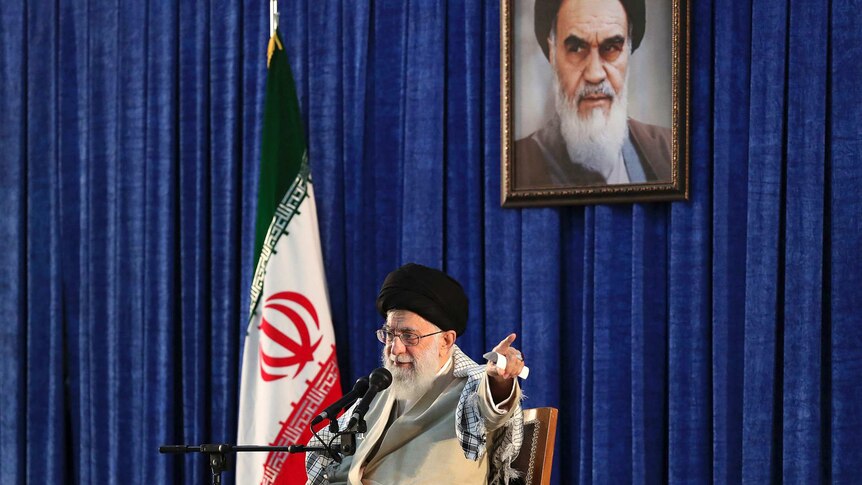 Iran's leader Ayotollah Khameini sits in front of a microphone, with an Iranian flag behind him.