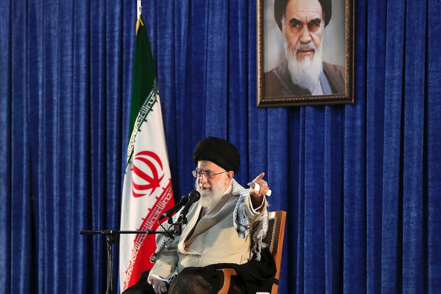 Iran's leader Ayotollah Khameini sits in front of a microphone, with an Iranian flag behind him.