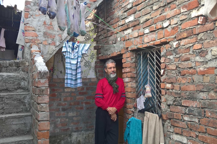Man with facial hair in red jumper gazes into the distance in a slum area. Clothes on washing line hang in the background.