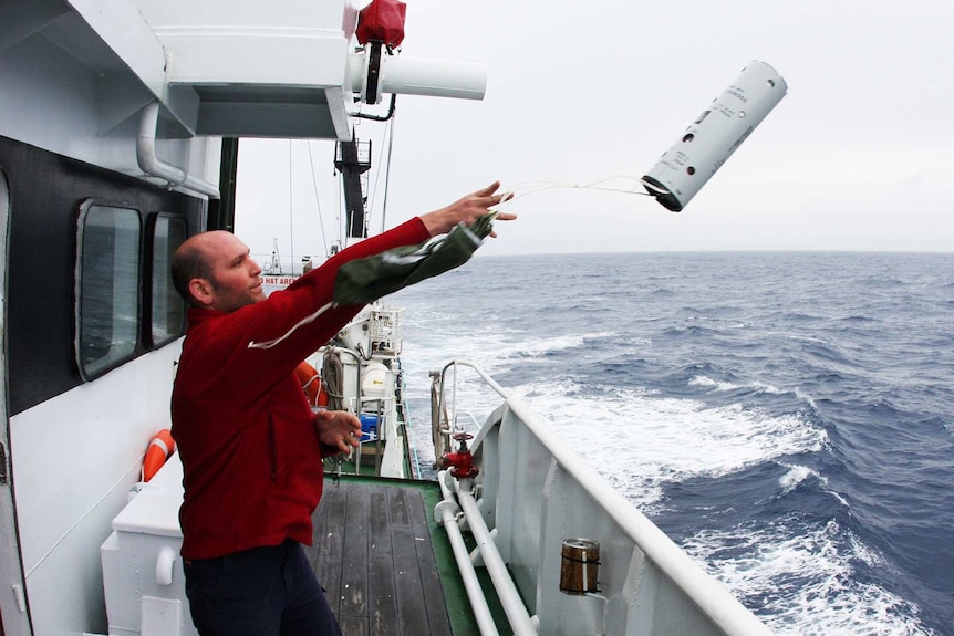 A man wearing a red jumper throws a device up into the air and off the deck of a boat.