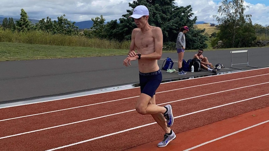 A man in a white cap and shirtless runs while checking his watch on a running track