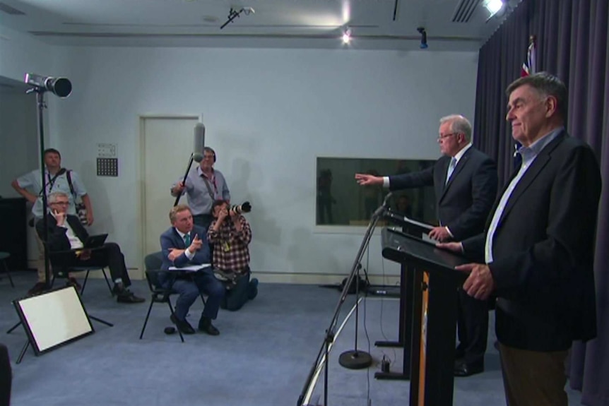 Wide shot press conference with Probyn trying to ask a question and Morrison standing at podium pointing to another journalist.