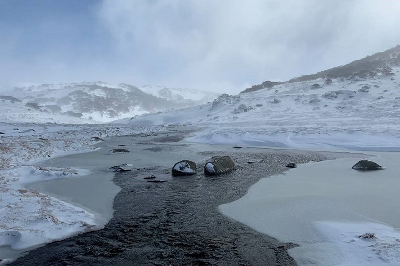 A snowy alpine waterway with a stream running and rocks in the foreground