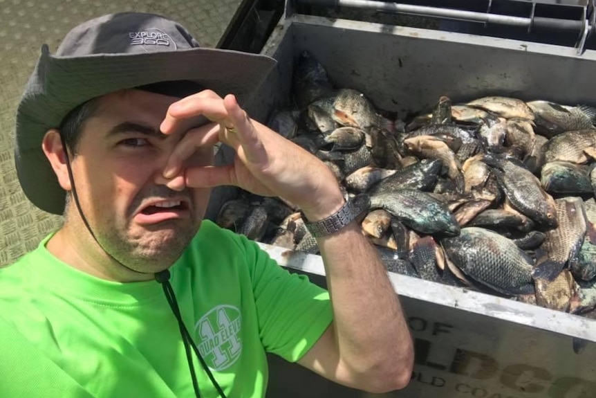 A man holds his nose near a skip bin full of dead fish.