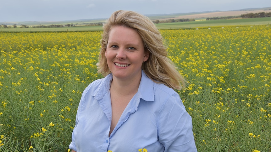 A woman with blonde hair, wearing a light blue shirt, standing in a field of canola.