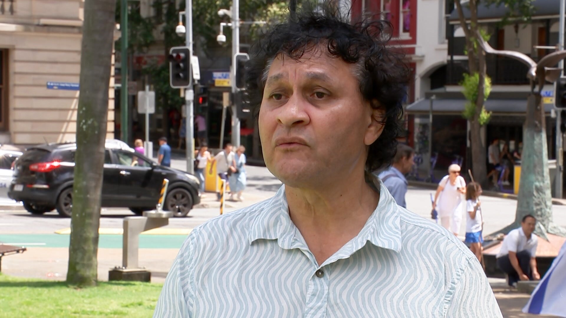a man wearing a white and light-blue shirt speaking to a reporter in a city square