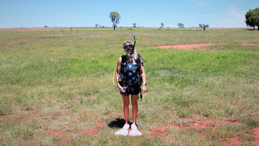 A woman in scuba diving equipment including snorkel and flippers stands in a paddock with red dirt and grass.