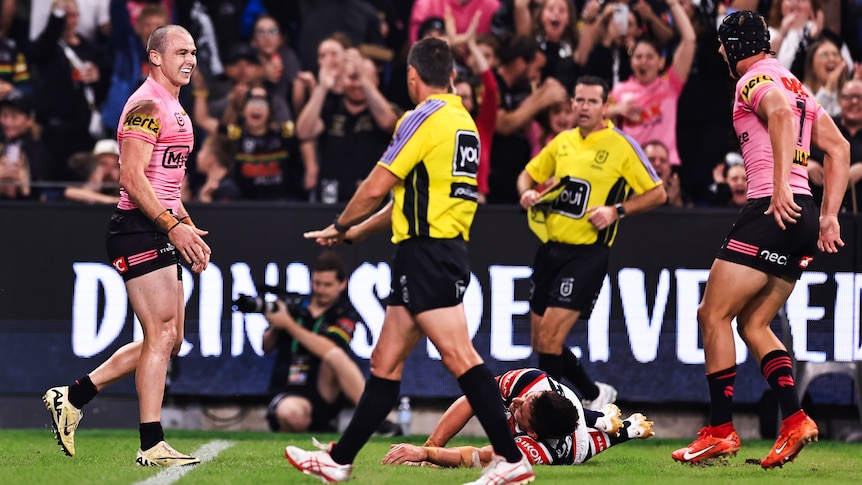 A Penrith NRL player grins as he runs back after scoring a try, as the referees stand next to a Roosters player on the ground.
