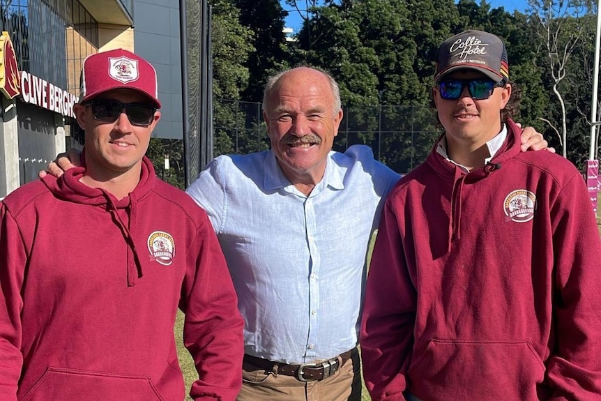 Two young soccer players in brown sweaters stand next to rugby league legend Wally Lewis, a man with white hair
