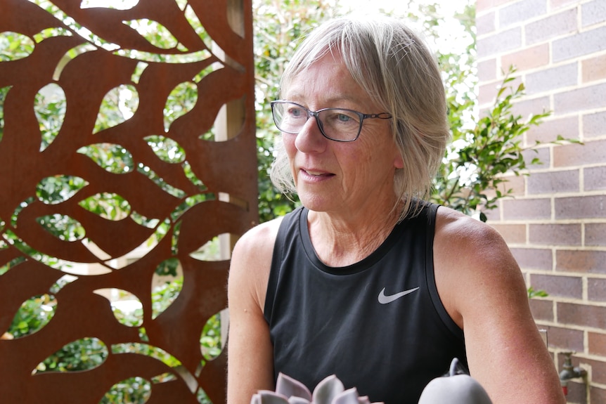A woman with grey hair and glasses looks off camera in a green space near a brick wall and a decorative metal screen.