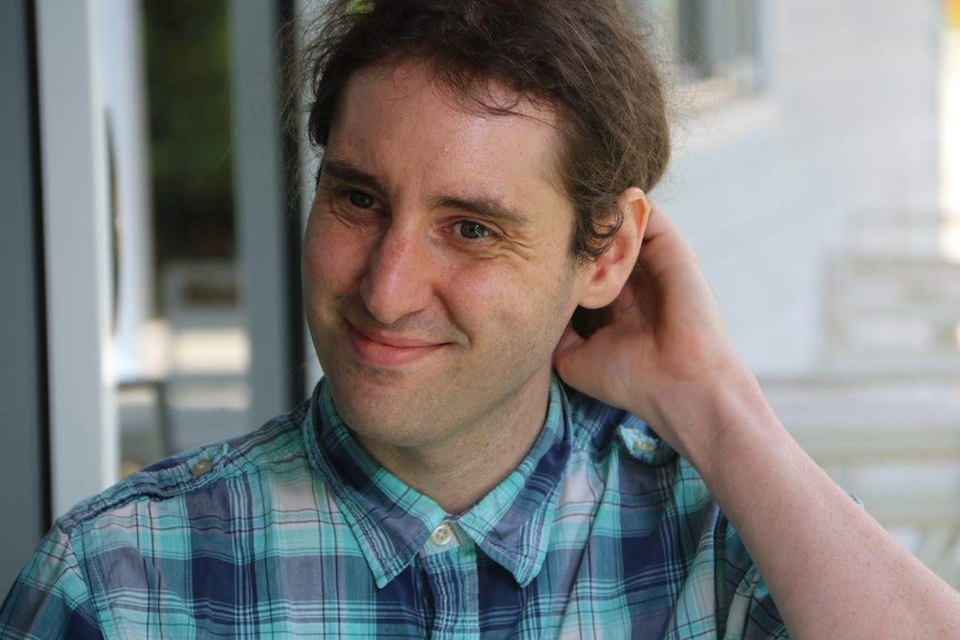 A young man with dark hair in blue shirt scratches the back of his head in an outdoor setting.