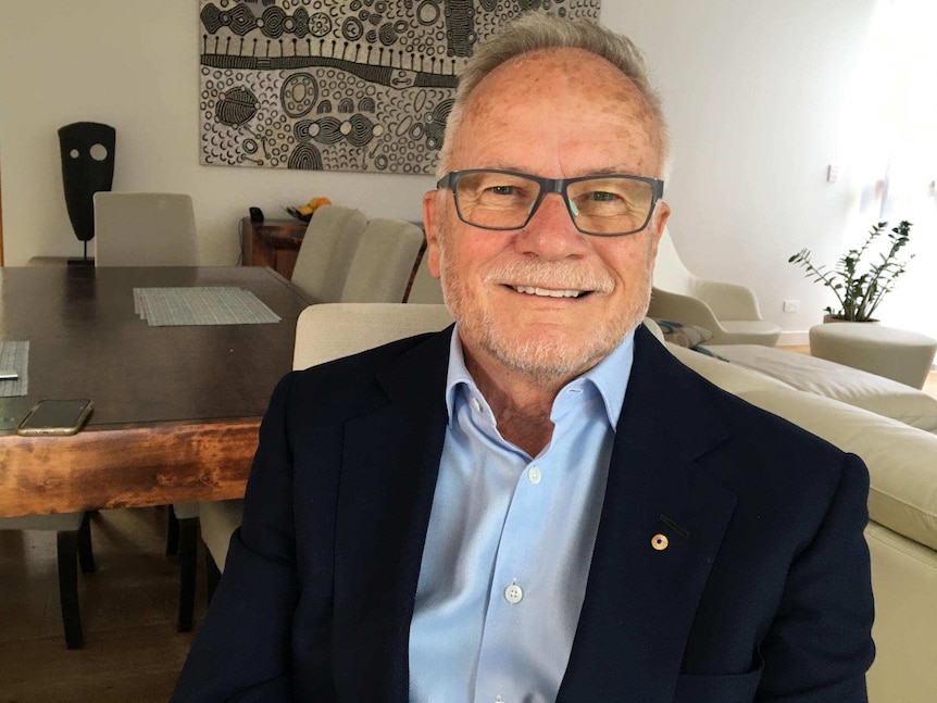 An elderly man wearing a light blue shirt and dark blue blazer smiles to the camera