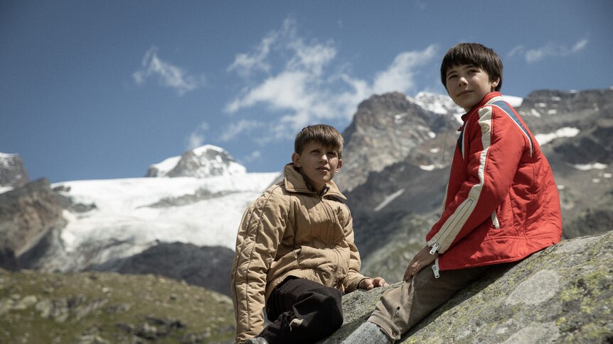 A young blonde Italian boy and a brunette Italian boy sit on a grassy hillside with snow-capped alps in the background.