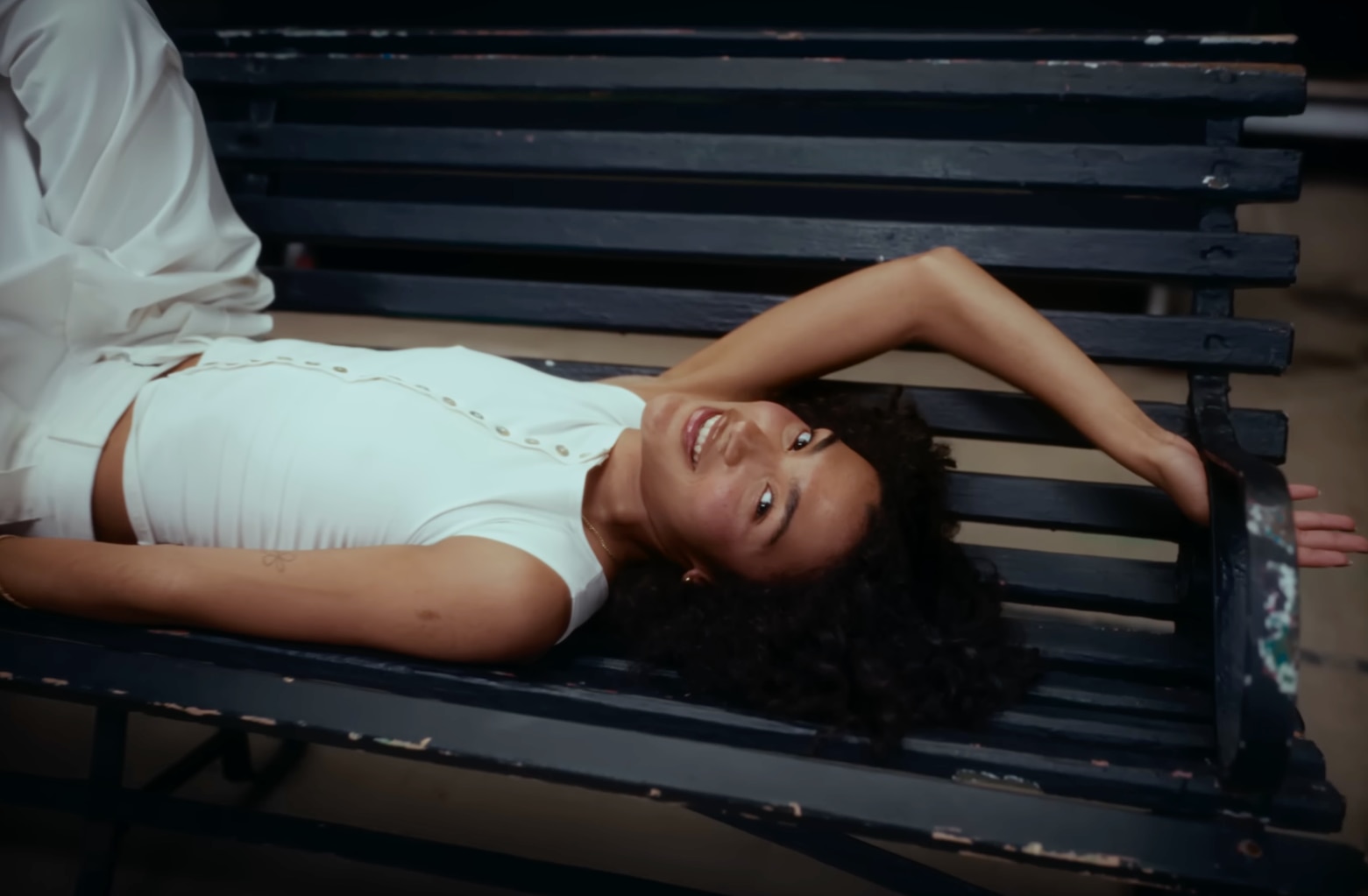 Olivia Dean yace en un banco negro del parque con un conjunto blanco sin mangas.