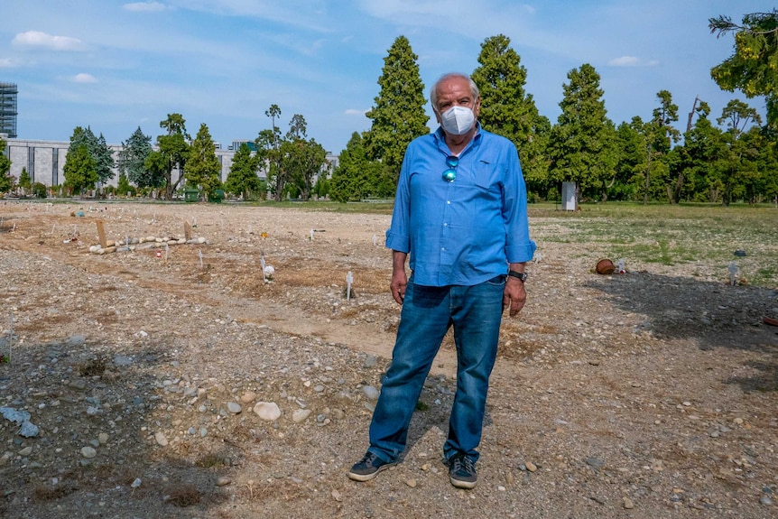 A man in a blue shirt and jeans and face mask stands in a grave yard