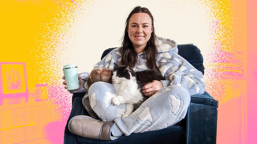 Ellen burns sits on a couch with a cat on her lap, wearing ugg boots, a fluffy blue onesie and holding a mug. 