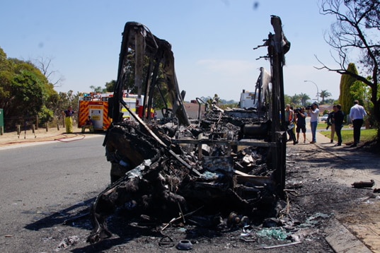 A front-on view of a bus sitting on the side of the road destroyed by fire, with the roof completely gone.