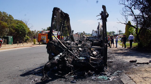 A front-on view of a bus sitting on the side of the road destroyed by fire, with the roof completely gone.