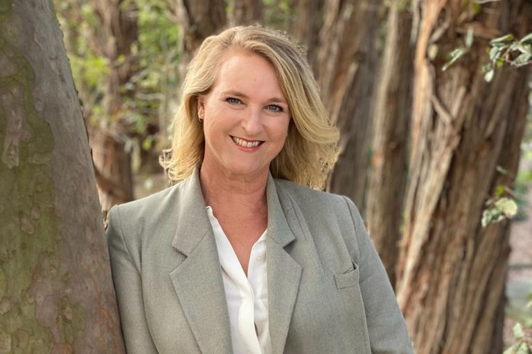 A woman with short blonde hair poses for a photo leaning against a tree trunk