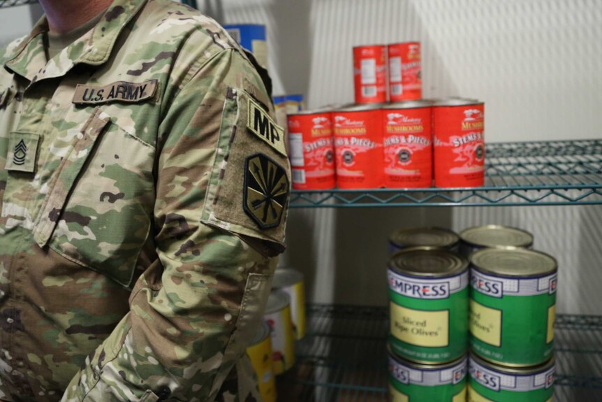 View of an unidentified soldier's torso standing in front of cans of food on a wire rack.