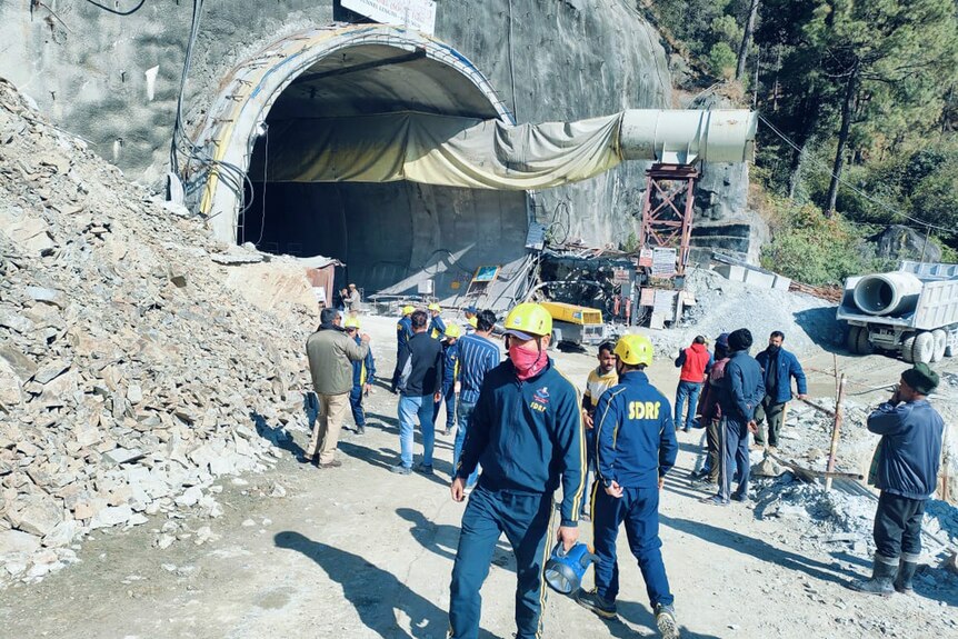 The building site of a tunnel is pictured with workers and rescue workers walking in front of it. 