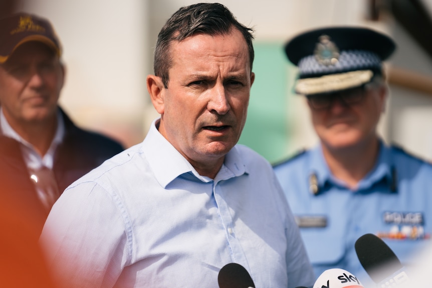The Premier in a light blue shirt speaks to press with the Police Commissioner in the background