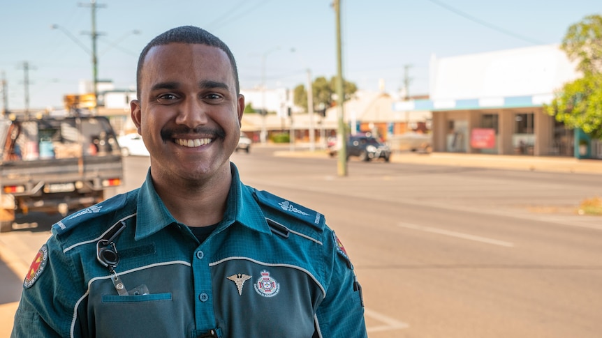 A young Indigenous man in a paramedic uniform stands outside, smiling.