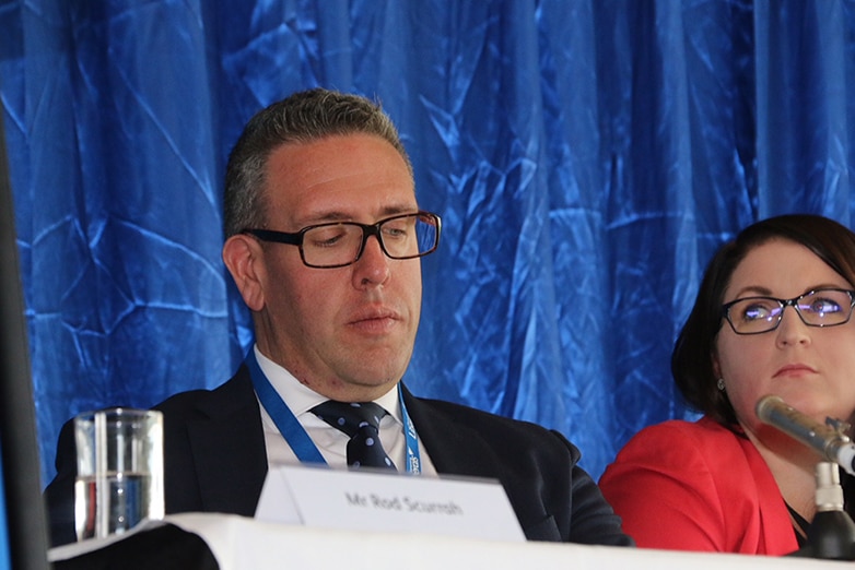 Sam McQuestin sitting at a table on a panel looking down.