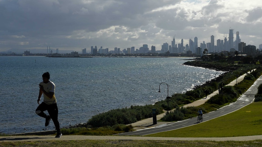 A silhouetted jogger climbs a hill with a city skyline, river path and cloudy skies behind.