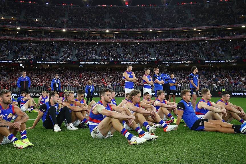 The losing team gathers together sitting on the ground as they watch the winners receive their AFL premiership medals.