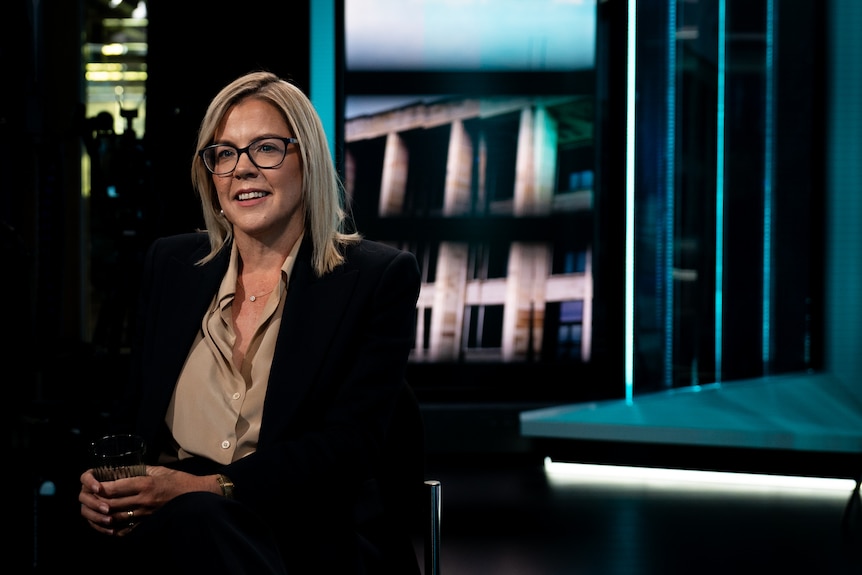 A mid shot of Libby Mettam sitting in a television studio. She is holding a glass of water and smiling.