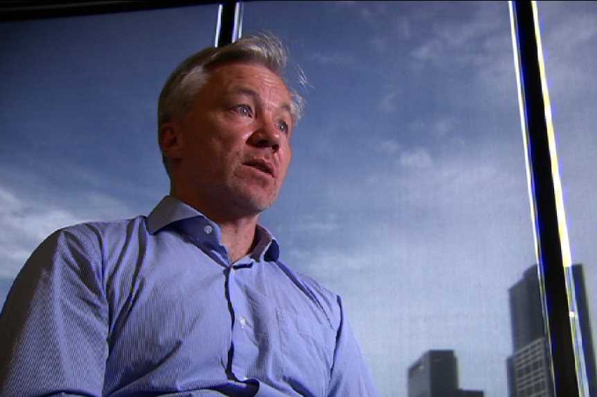 A grey-haired man in a business shirt stands in front of an office window.