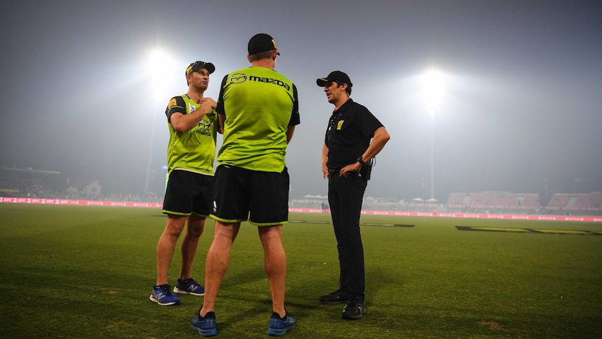 Officials stand around as floodlights illuminate the thick smoke haze at Manuka Oval