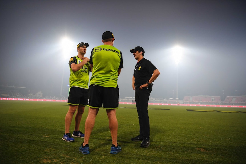 Officials stand around as floodlights illuminate the thick smoke haze at Manuka Oval