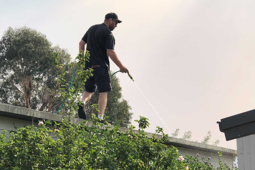 A man stands on a roof using a water hose to clear gutters.