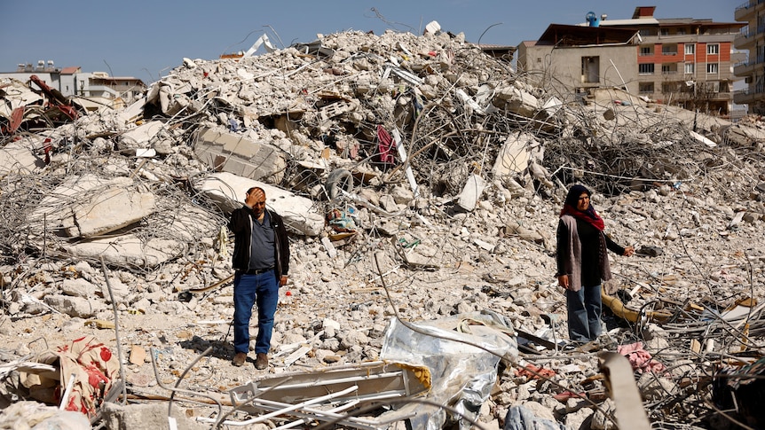 A man and woman stand among rubble. 