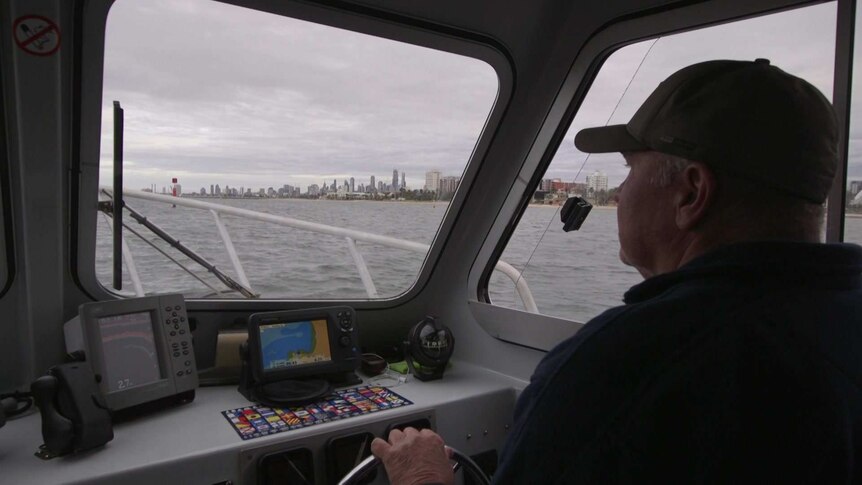 A man wearing a hat steers a boat, with the Melbourne skyline in view over the water.