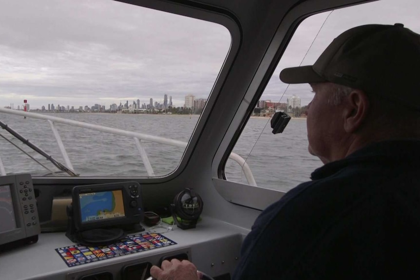 A man wearing a hat steers a boat, with the Melbourne skyline in view over the water.